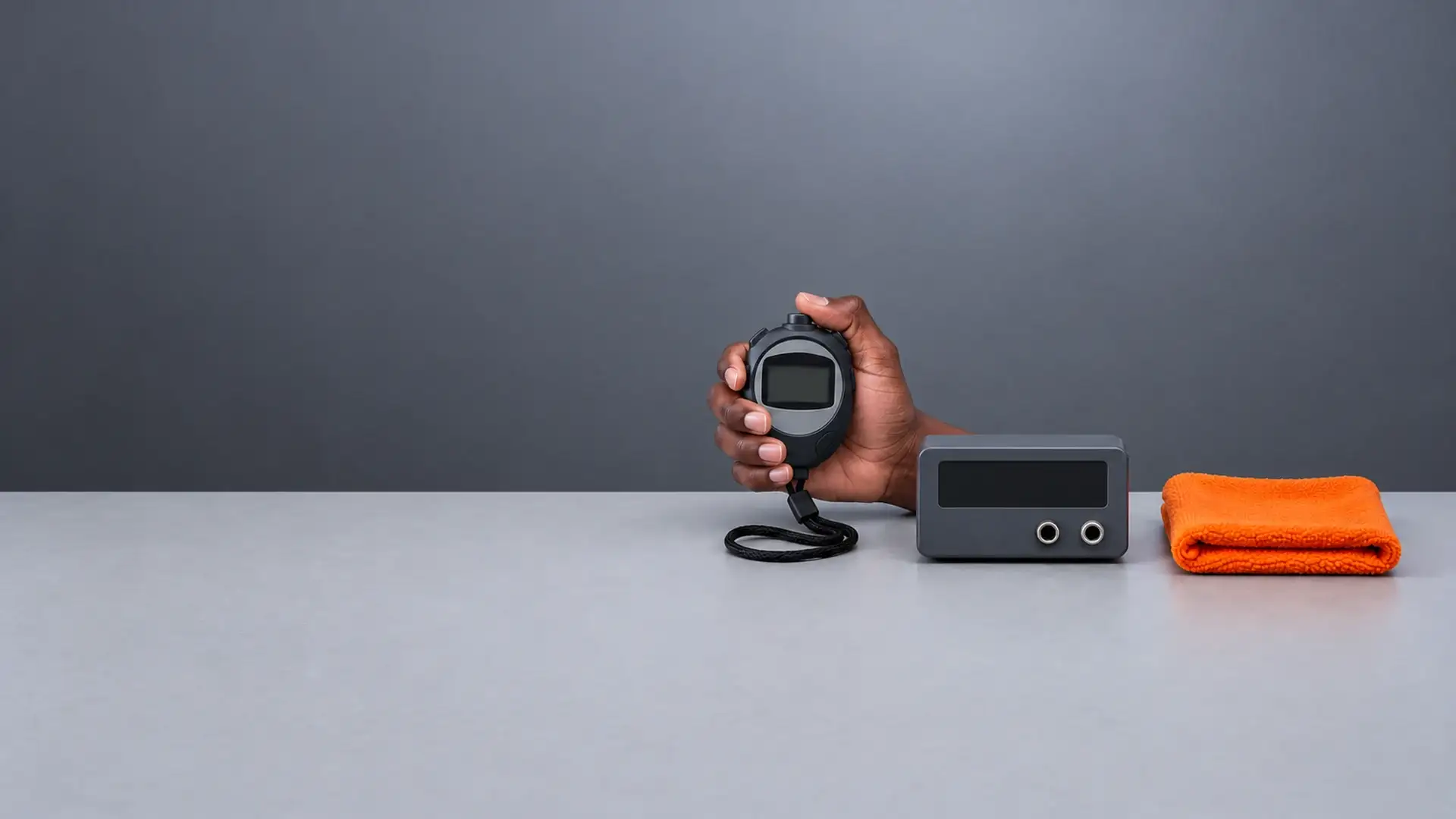 Mão segura um cronômetro digital de mão, com o cordão pendurado, ao lado de um padrão de tempo de bancada