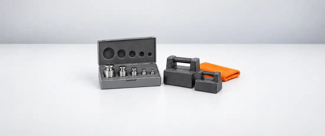 Estojo aberto com cinco massas-padrão de inox, ao lado de duas massas de ferro fundido com alça, na bancada