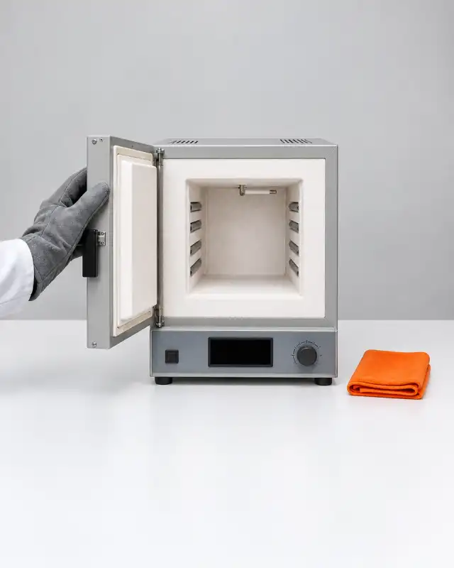 Câmara branca e fria de uma mufla de porta aberta, com as resistências nas laterais e a porta apoiada por mão de luva térmica